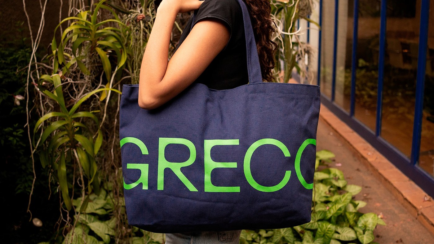 Screenprinted canvas tote bag with bold Grecotipo typography carried in outdoor studio setting