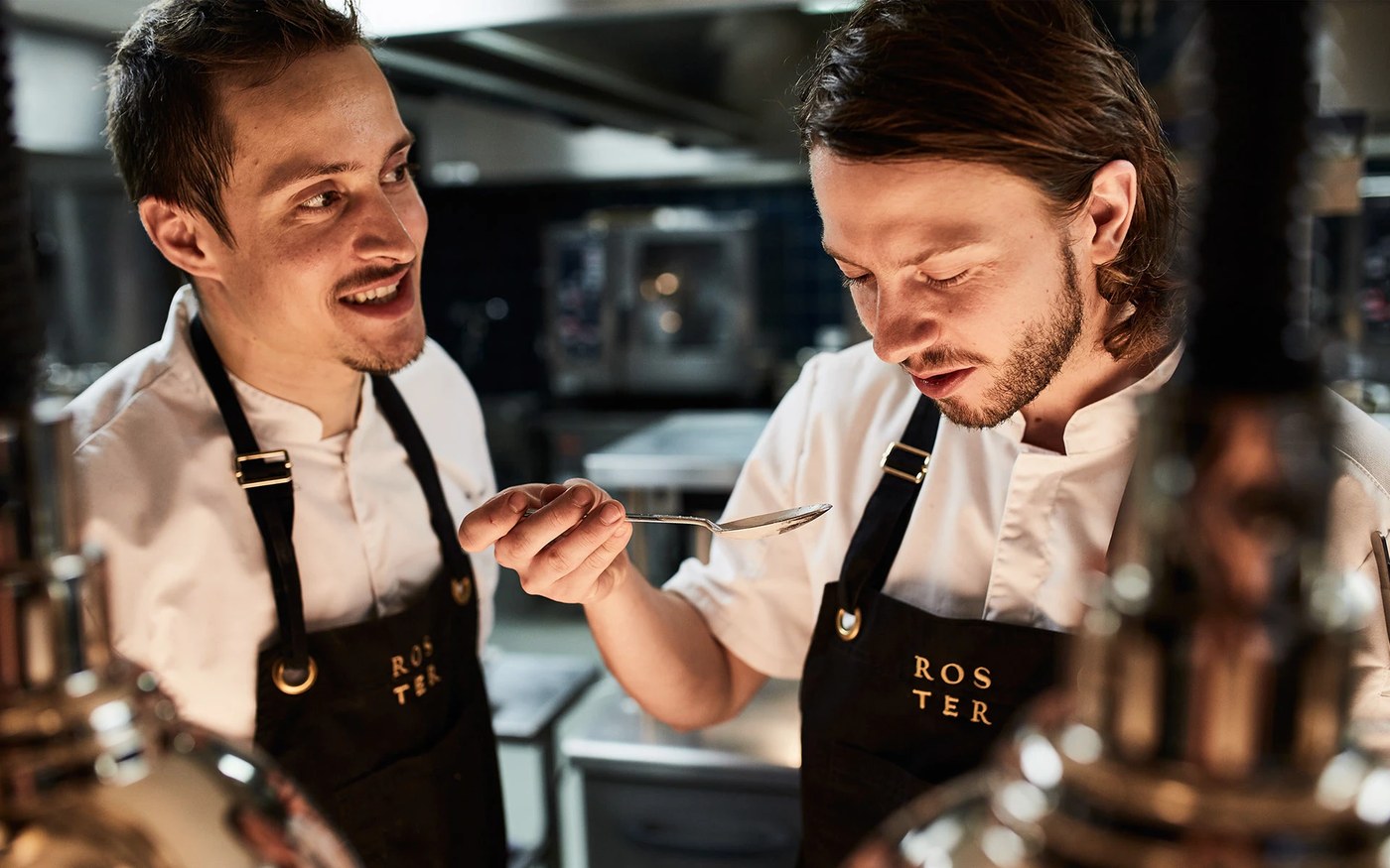 Founders Kari Aihinen and Paul Hickman wearing embroidered black canvas aprons in the kitchen for Roster branding design