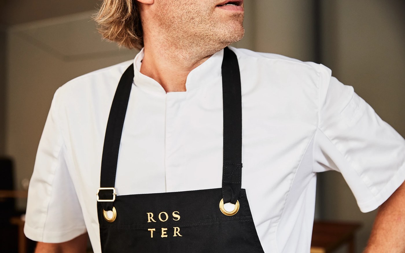 Close-up detail of black canvas kitchen apron with brass grommets and gold embroidered wordmark in Roster branding design